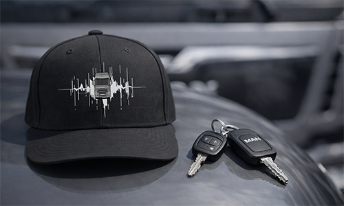 Black cap with truck and soundwave design, placed on reflective surface with MAN vehicle keys beside