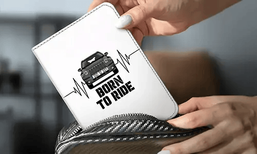Person holding white wallet with black car and heartbeat design, bold BORN TO RIDE text, carbon-fiber pouch in background