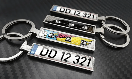 Schlüsselanhänger mit Nummernschild im Comic-Stil, angeordnet auf einer carbonstrukturierten Oberfläche mit farbenfrohen Autografiken