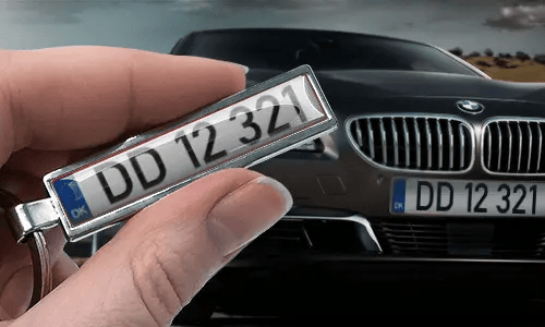 Portachiavi a forma di targa in stile fumetto con numero personalizzato, tenuto in mano vicino alla targa dell’auto corrispondente